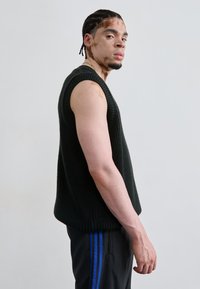 Mørkegrøn strikket ærmeløs sweater med et mønster i tekstur, båret over sorte bukser med blå striber. Modellen står i profil.