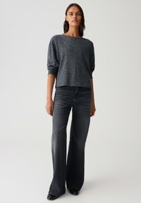 Femme debout portant un pull ample gris foncé et un jean large taille haute délavé noir avec des chaussures noires contre un fond uni.