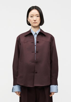 Mujer con cabello liso y negro que lleva una chaqueta granate abotonada sobre una camisa azul a rayas con puños extendidos, de pie frente a un fondo liso.