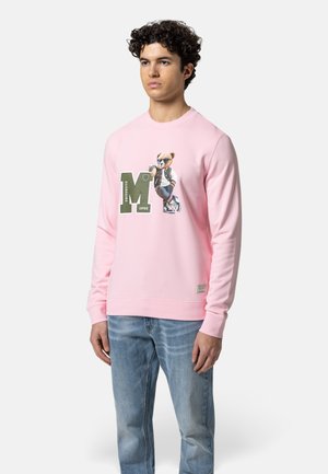 Mann trägt rosa Sweatshirt mit grünem "M" und Bärenmotiv, kombiniert mit hellblauen Jeans, steht mit entspannten Armen.