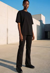 Person wearing black oversized T-shirt, black pants, and black sneakers standing on concrete near modern minimalistic building under clear blue sky.