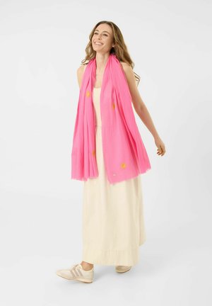 Vrouw glimlacht, draagt een lange beige jurk, witte sneakers en een felroze sjaal met gele zonpatronen, staand op een witte achtergrond.