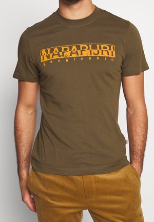 Mężczyzna w oliwkowozielonym t-shircie z krótkim rękawem z pomarańczowym logo "Napapijri geographic" oraz musztardowych mankietowych spodniach, ręce w kieszeniach.