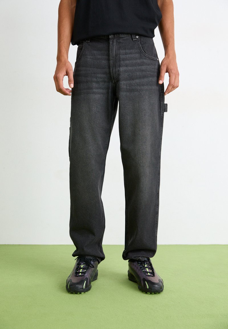 Pantalons cargo en denim noir avec une coupe droite, présentant des poches latérales, un léger délavage et des coutures contrastées. Associés à des baskets noires.
