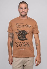 T-shirt en coton marron avec un imprimé graphique noir d'un casque avec des ailes et le texte en gras "THUNDER RACE", col rond, manches courtes et coupe décontractée.