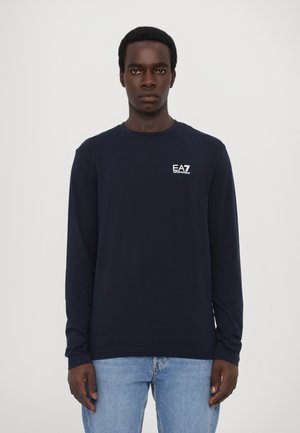 Long-sleeve navy T-shirt made of cotton with a crew neck and EA7 logo printed in white on the left chest. Worn with light blue jeans.