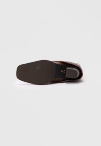 Blick von unten auf einen braunen Lederschuh mit glattem Profil und rundem Zehenbereich, ausgestattet mit einer strukturierten Gummisohle und einem goldenen Akzent-Logo.