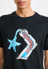 Camiseta negra con un estampado gráfico de un líquido azul vertiéndose de un tubo con "CONVERSE" en letras en negrita rojas y azules.