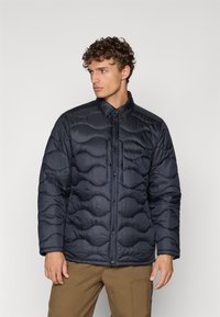 Marinblå puffjacka med kviltad design, tryckknappar och krage. Har en strukturerad yta och en bröstficka fram.