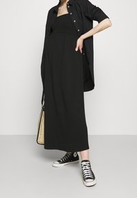 Raska koja nosi dugu crnu haljinu bez naramenica, preveliku crnu košulju na dugmad, crne tenisice na visokom potpetici i nosi bež pleteni ranac.