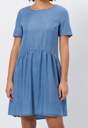 Frau trägt ein hellblaues, kurzärmeliges Jeanskleid mit geraffter Taille und rundem Ausschnitt vor einem schlichten hellen Hintergrund.