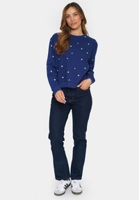 Felpa navy con motivi a stelle bianche, collo rotondo e polsini a costine, abbinata a jeans scuri e sneakers grigie.