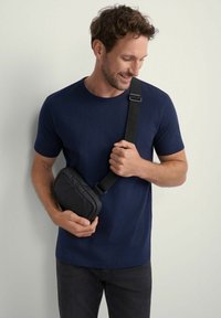 Czarna torba na ramię z wypełnieniem, noszona na granatowej koszulce. Widać regulowany pasek oraz zamek błyskawiczny. Gładka faktura.