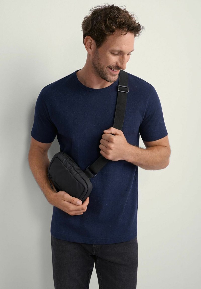 Czarna torba na ramię z wypełnieniem, noszona na granatowej koszulce. Widać regulowany pasek oraz zamek błyskawiczny. Gładka faktura.