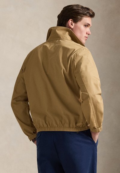 Καφέ μπουφάν bomber με ψηλό γιακά, ελαστικό τελείωμα και μανσέτες. Διαθέτει λεία υφή και άνετη εφαρμογή, φορεμένο πάνω από σκούρο παντελόνι.