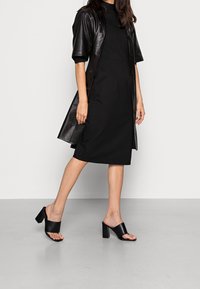 Robe noire en tissu lisse avec des manches courtes en cuir, présentant un corps ajusté et une jupe droite. Associée à des sandales noires à talons carrés.