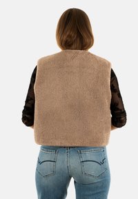 Gilet in pile beige con superficie texturizzata, senza maniche e colletto rotondo, indossato sopra una maglietta nera a maniche lunghe e jeans azzurri chiari.