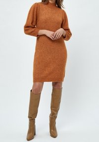 Femme portant une robe en maille côtelée de couleur rouille avec manches longues et des bottes à talons hautes jusqu'aux genoux de couleur fauve, se tenant devant un fond uni.