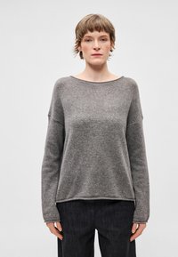 Szary sweter z dzianiny o szerokim dekolcie, długich rękawach i luźnym kroju. Miękka tekstura i lekko zaokrąglony dół. Zestawiony z ciemnymi spodniami.