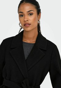 Manteau en laine noir avec un col, une taille ceinturée et un tissu texturé. Porté par-dessus un haut gris, il dispose de fermetures à boutons et de poches latérales.