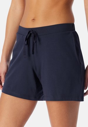 Frau trägt dunkelblaue Shorts mit Kordelzug und Seitentaschen, zeigt Bauchbereich und Oberschenkel vor einfarbigem Hintergrund.