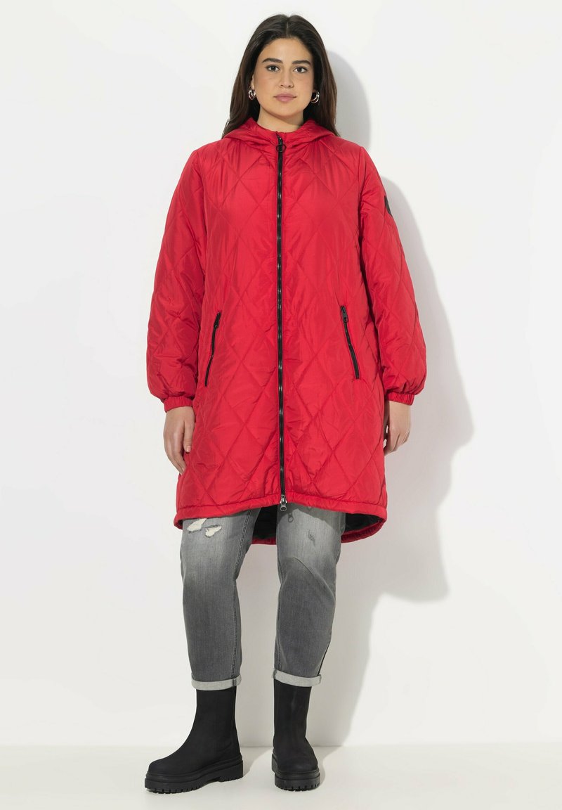 Red, quilted long jacket with a zipper front, elastic cuffs, and two side pockets. Worn over gray distressed jeans and black ankle boots.