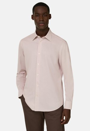 Homme portant une chemise rose clair à boutons avec manches longues et un pantalon marron foncé, debout devant un fond blanc uni.