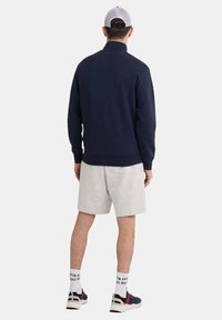 Marineblå pullover med høj krave, ribbede manchetter, parret med grå shorts, hvide sokker og sporty sneakers, set fra ryggen.