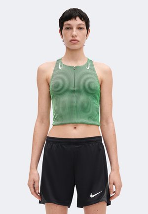 Γυναίκα που φορεί πράσινο ριγέ αμάνικο crop top και μαύρο αθλητικό σορτς Nike, στέκεται απέναντι με ουδέτερη έκφραση.