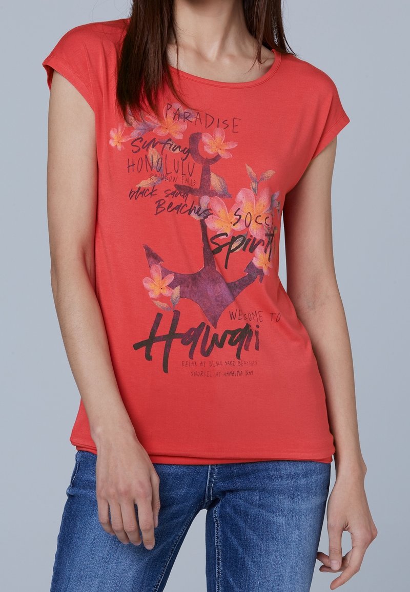 T-shirt rouge à manches courtes en coton doux, avec un motif floral, du texte sur le thème des vacances et une coupe décontractée. Associé à un jean en denim.
