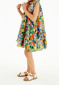 Ärmelloses Kleid mit Blumenmuster in leuchtenden Farben und rüschigem Ausschnitt, kombiniert mit weißen Sandalen mit Riemen und brauner Sohle.