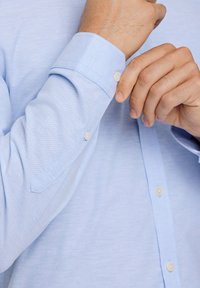 Světle modrá košile s dlouhými rukávy a hladkou texturou, s špičatými manžetami a malými bílými knoflíky. Doplněná kontrastním šitím.