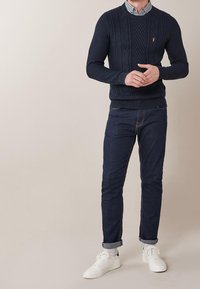 Granatowy sweter w warkocz z okrągłym dekoltem; noszony na jasnej koszuli w kratkę, w połączeniu z ciemnymi dżinsami i białymi trampkami.