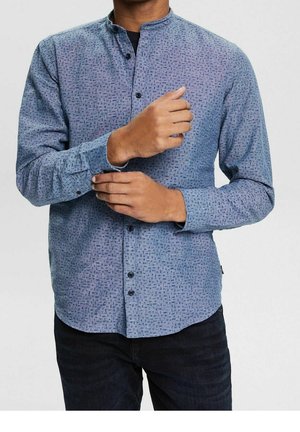 Mand, der justerer manchet på blå mønstret skjorte med mandarinkrave, iført mørke bukser, mod en ensartet lys baggrund.