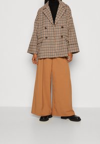 Giacca doppiopetto a quadri nei toni crema, marrone e nero, con maniche ampie, abbinata a pantaloni beige a gamba larga e stivali neri con tacco a blocco.