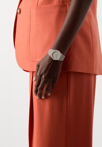 Montre grise avec un cadran rond sur un bracelet texturé, assortie à un costume en tissu corail avec une grande poche et des lignes épurées.
