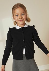 Svart stickad cardigan med rund hals, knappstängning fram och volangdetaljer på axlarna. Bärs över en vit skjorta med krage.
