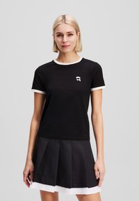 Black short-sleeve t-shirt with white trim, featuring a small graphic logo. Paired with a pleated black skirt with a white underlayer.
