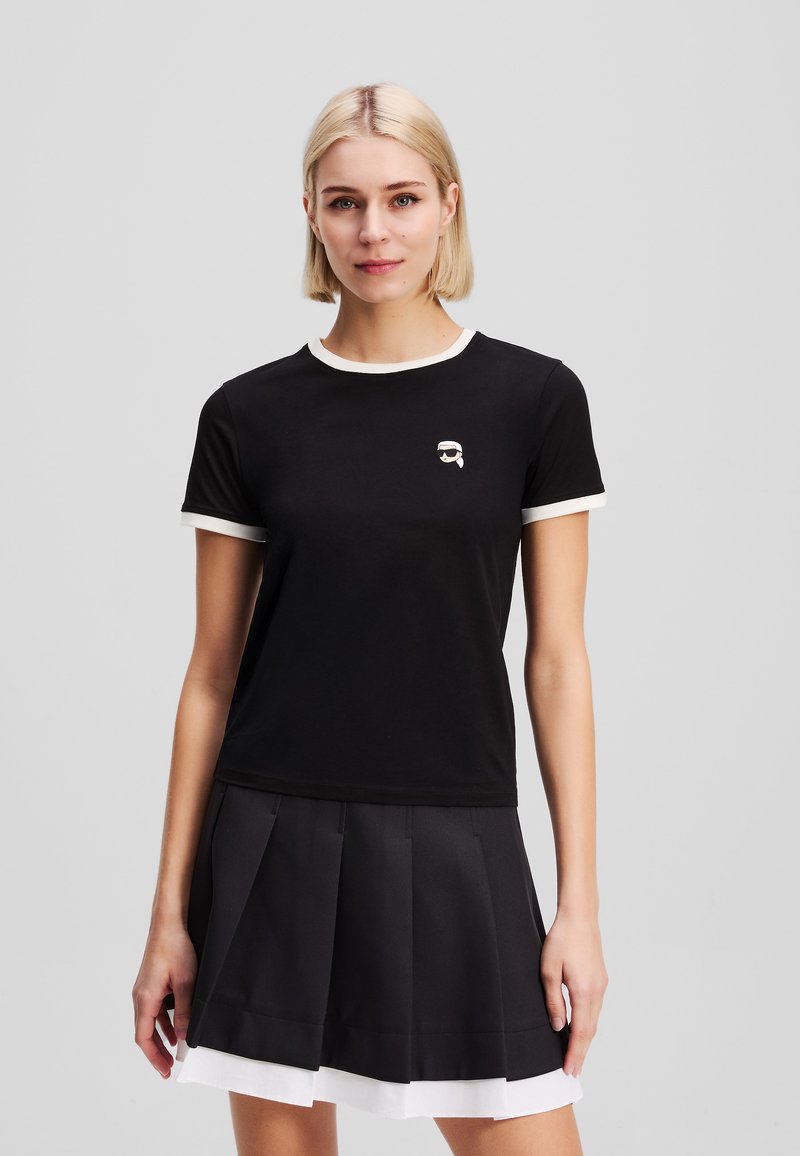 Black short-sleeve t-shirt with white trim, featuring a small graphic logo. Paired with a pleated black skirt with a white underlayer.