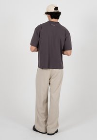 Homme portant une casquette beige, un t-shirt gris foncé, un pantalon beige et des chaussures noires, debout de dos face à la caméra sur un fond uni.