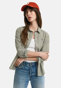 Striped green and white shirt with long sleeves, button closure, worn over a white tank top and paired with faded blue jeans. Red cap on top.