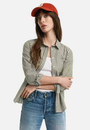 Grün-weiße gestreifte Bluse mit langen Ärmeln, Knopfverschluss, über einem weißen Tanktop getragen und mit verblassten blauen Jeans kombiniert. Rote Kappe auf dem Kopf.