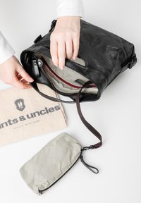 Sac à épaule en cuir noir avec un intérieur rayé, une fermeture éclair rouge contrastante et une petite pochette grise attachée, reposant sur un sac beige.