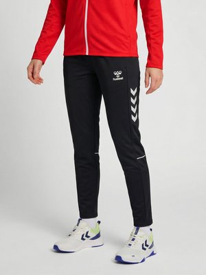 Persoon die zwarte hummel trainingsbroek draagt met witte chevronlogo's, een rood jasje met ritssluiting, witte sokken en witte sneakers met blauwe accenten.
