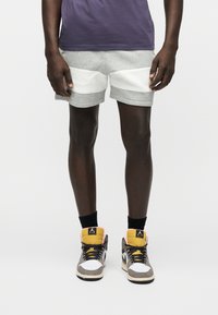 Άνδρας με γκρι σορτς με λευκές επιφάνειες, μαύρες κάλτσες και πολύχρωμα ψηλά αθλητικά παπούτσια, στέκεται μπροστά σε λευκό φόντο.