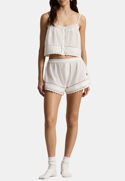 Woman wearing white sleeveless crop top and matching elastic waist shorts with lace trim and white socks on a plain background.