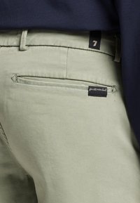 Lys grøn chinos med en glat tekstur, som har en baglomme og et sort mærke. Livremmen har et bælte og syninger.