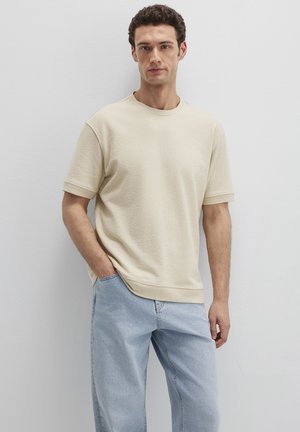 Mann trägt beige kurzärmliges Hemd und hellblaue Jeans, steht mit einer Hand in der Tasche vor einer schlichten weißen Wand.