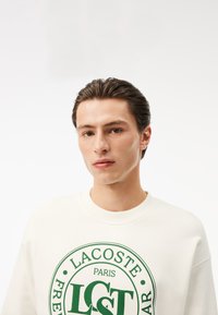 Felpa bianca con logo verde rotondo, con la scritta "LACOSTE PARIS" e "LCST." Texture liscia, design classico con collo a giro.