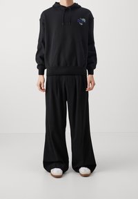 Black hoodie with a front pocket and graphic text, paired with loose-fitting black trousers. Shoes are white with brown soles.